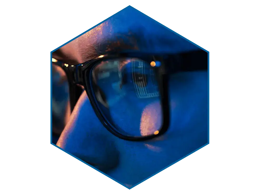 The left eye of a person wearing dark-rimmed glasses, with the faint reflection of a computer screen in the lens